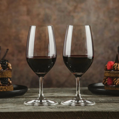 Two wine glasses filled with red wine on a wooden table with desserts in the background.
