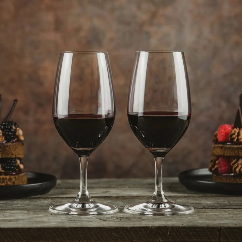 Two wine glasses filled with red wine on a wooden table with desserts in the background.